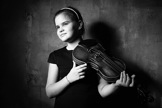 A young girl with a violin stands in front of a wall.  