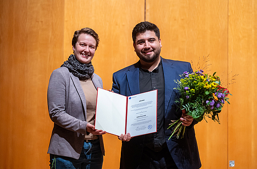 The picture shows two people with a certificate and flowers.