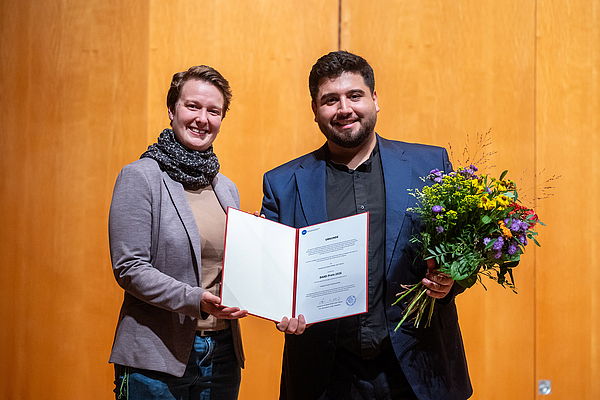 Das Bild zeigt zwei Menschen mit einer Urkunde und Blumen.