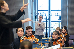 Das Bild zeigt junge Menschen mit Instrumenten und eine Dirigentin.
