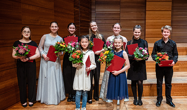 Das Bild zeigt neun junge Menschen, die mit Blumen und Urkunden in der Hand auf einer Bühne stehen. 