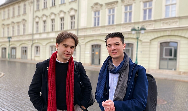 Zwei junge Männer stehen vor einem Gebäude, sie schauen in die Kamera