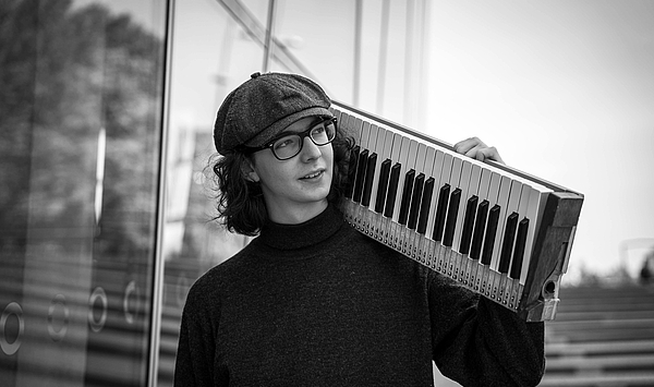 Das Bild zeigt einen jungen Mann mit Mütze, der ein Keyboard auf der Schulter trägt. Das Bild zeigt einen jungen Mann mit Mütze, der ein Keyboard auf der Schulter trägt.