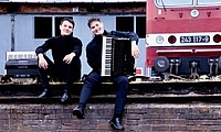 Zwei junge Männer mit Akkoredeons sitzen an einem Bahnhof