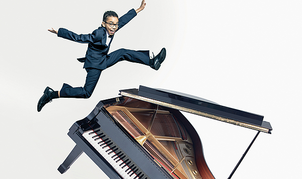Das Bild zeigt einen jungen Mann, der über einem Klavier springt. 