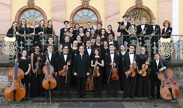 Pupils in concert dress stand in front of a castle building. They hold their instruments in their hands. Schüler und Schülerinnen stehen in Konzertkleidung vor einem Schlossgebäude. Sie halten ihre Instrumente in der Hand.