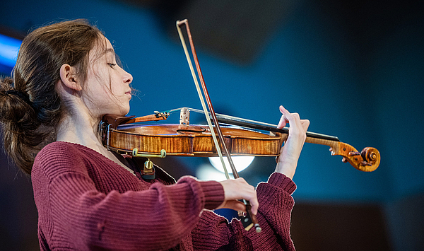 Das Bild zeigt eine junge Frau mit dunklen Haaren, die Geige spielt. 