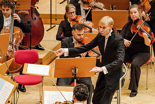 The picture shows a conductor in front of an orchestra.