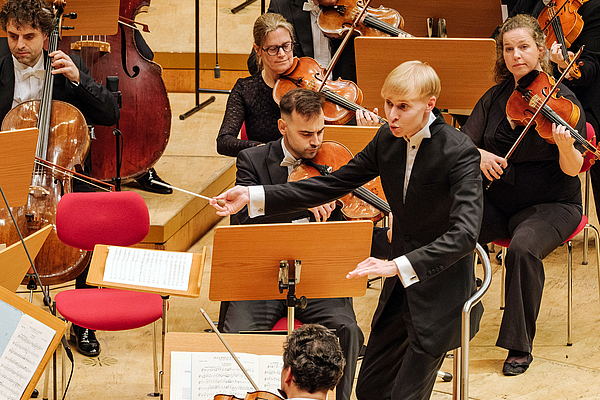 Das Bild zeigt einen Dirigenten vor einem Orchester.