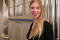 A young woman with blonde hair stands in front of organ pipes