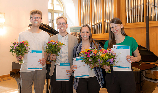 The picture shows four young people with certificates and flowers. Das Bild zeigt vier junge Menschen mit Urkunden und Blumen.
