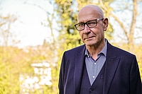 A man with glasses looks into the camera, trees can be seen in the background