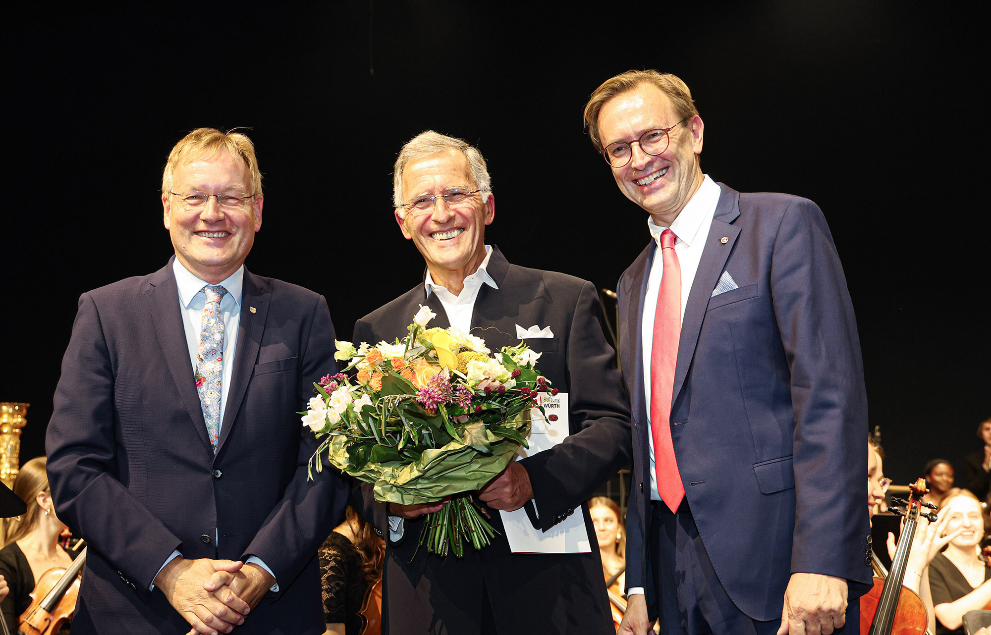Das Bild zeigt drei Personen und einen Blumenstrauß.