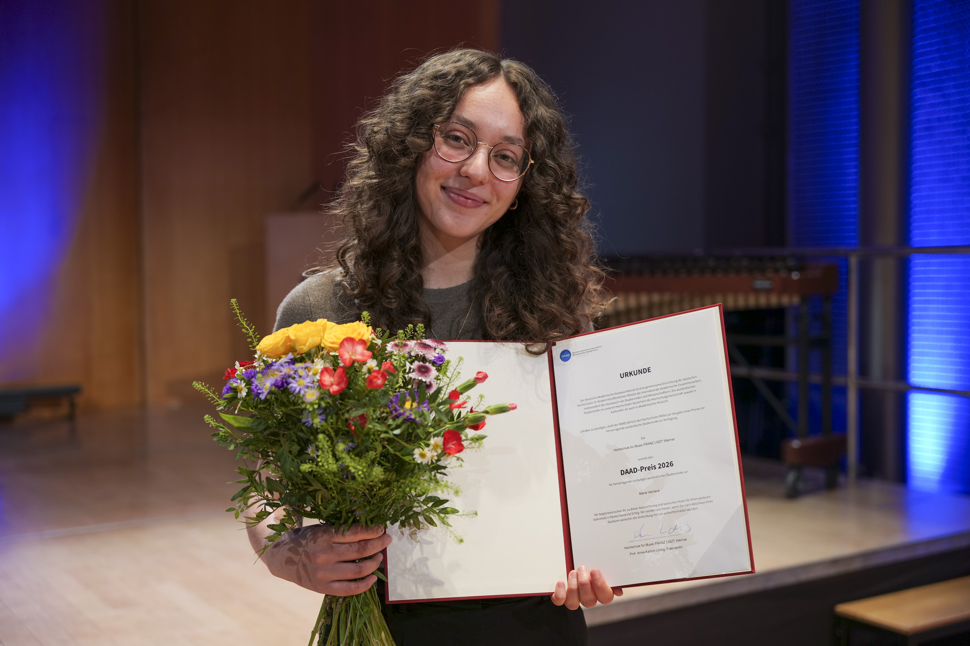 Das Bild zeigt eine Person mit Blumen und einer Urkunde.