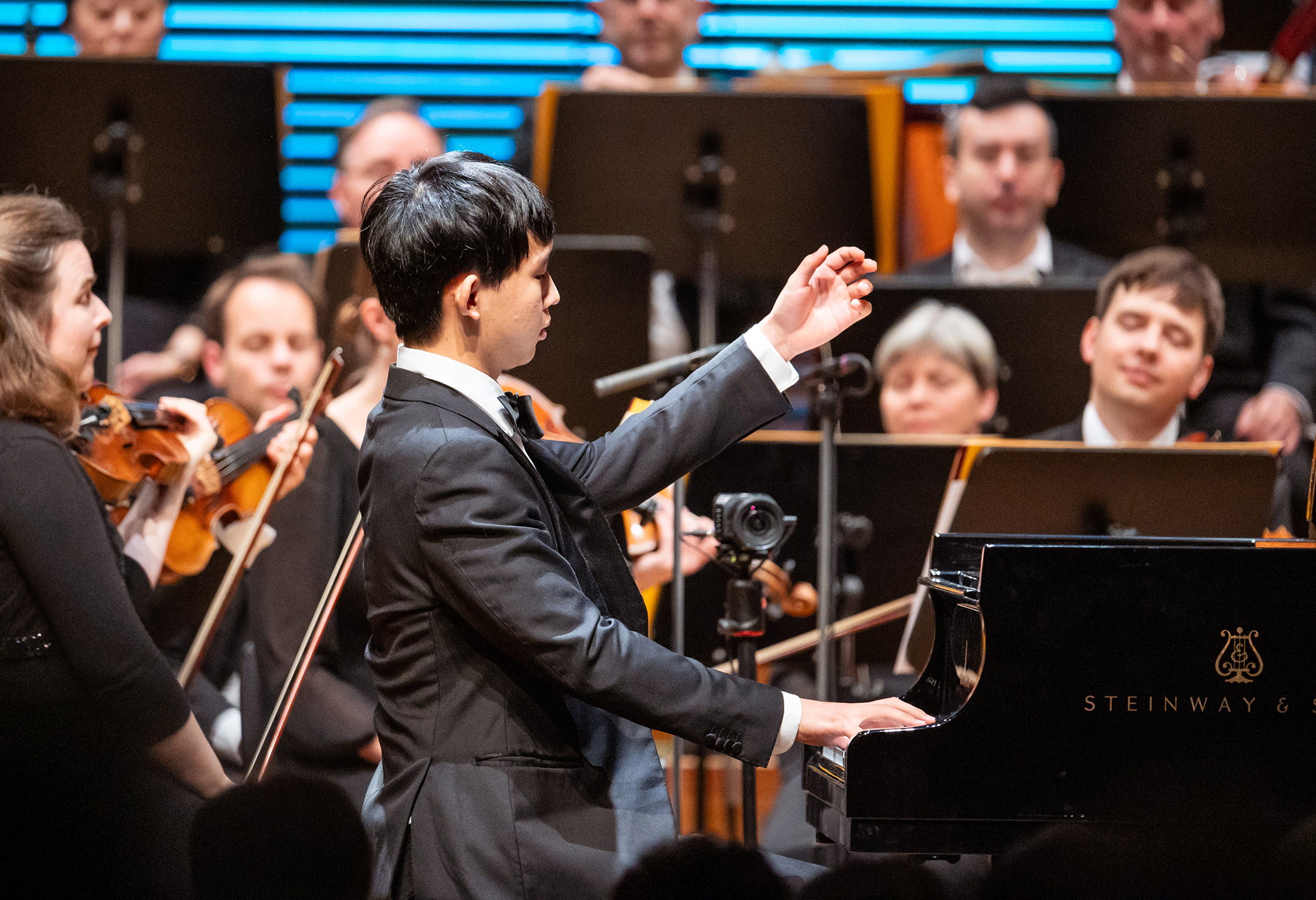 Das Bild zeigt eine Person an einem Konzertflügel vor einem Orchester.