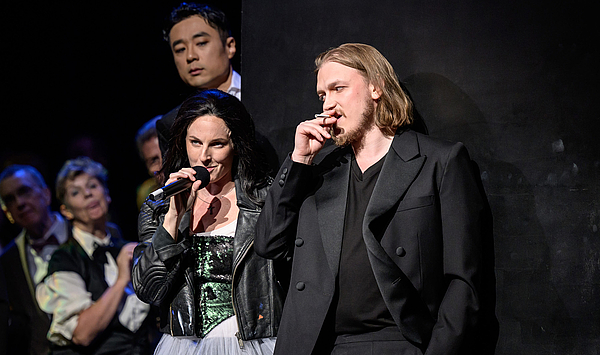 Das Bild zeigt einen Mann mit Zigarette und eine Frau mit Mikrophon.