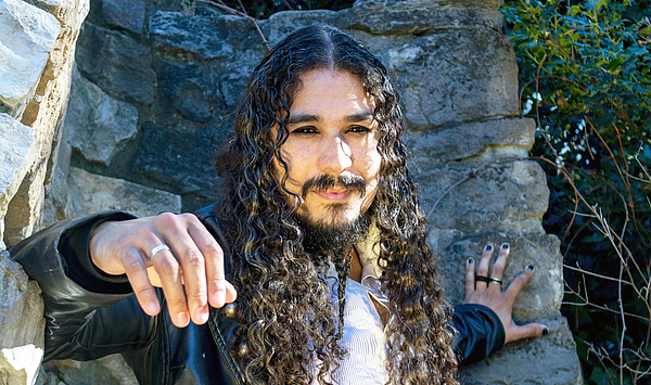 Ein Mann mit dunklen, lockigen Haaren lehnt an einem Felsen. 