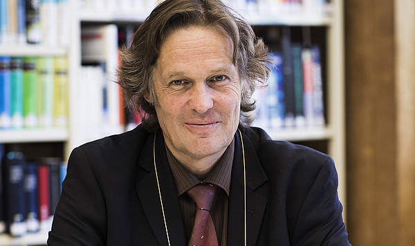 The picture shows a man in front of a bookshelf. Das Bild zeigt einen Mann vor einem Bücherregal.