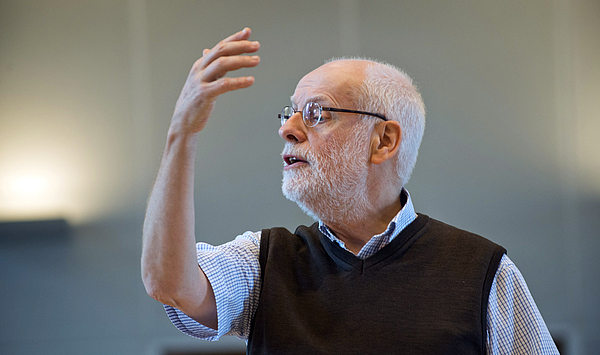A man with grey hair and a grey beard raises one arm. Ein Mann mit grauen Haaren und grauem Bart hebt einen Arm.