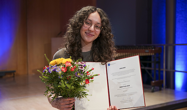 Das Bild zeigt eine Person mit Blumen und einer Urkunde.