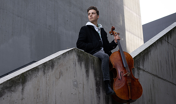 Das Bild zeigt einen Mann mit einem Cello auf einer Mauer.